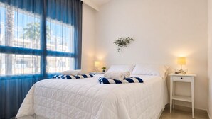 A neatly made bed with white bedding and blue striped pillows, a bedside table with a lamp, and a window with blue curtains.