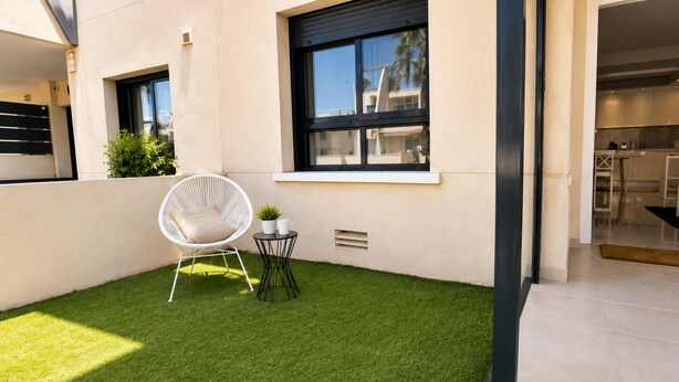A modern outdoor area with artificial grass, a white wicker chair, a small table with a plant, and a window.
