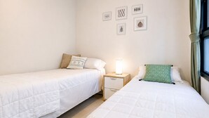 A hotel room with two single beds, a bedside table with a lamp, a green pillow, and wall art.