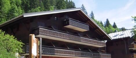 A two-story wooden chalet with balconies, surrounded by lush greenery and a forested backdrop.