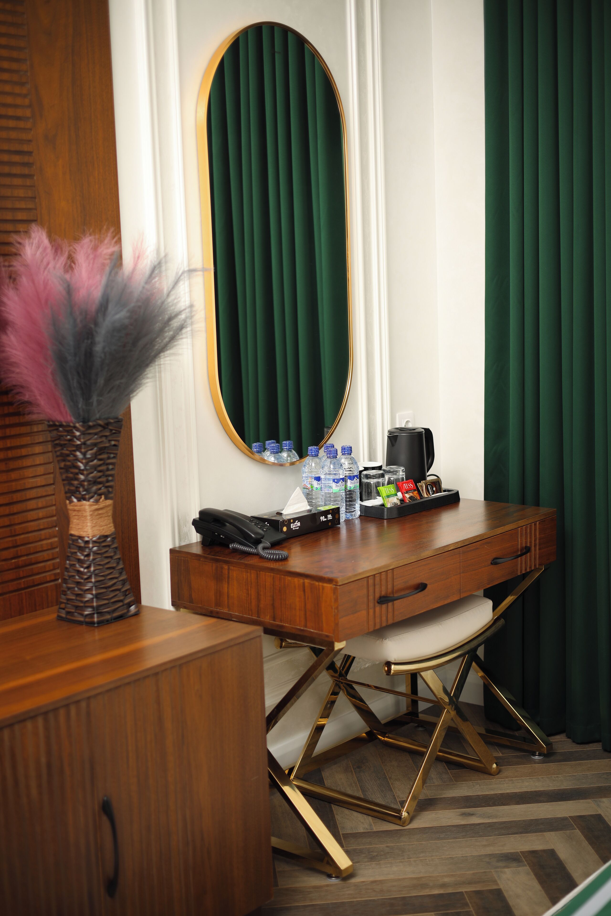 A hotel room with a wooden desk, a mirror, a vase with feathers, a telephone, a coffee maker, and a tray with toiletries.