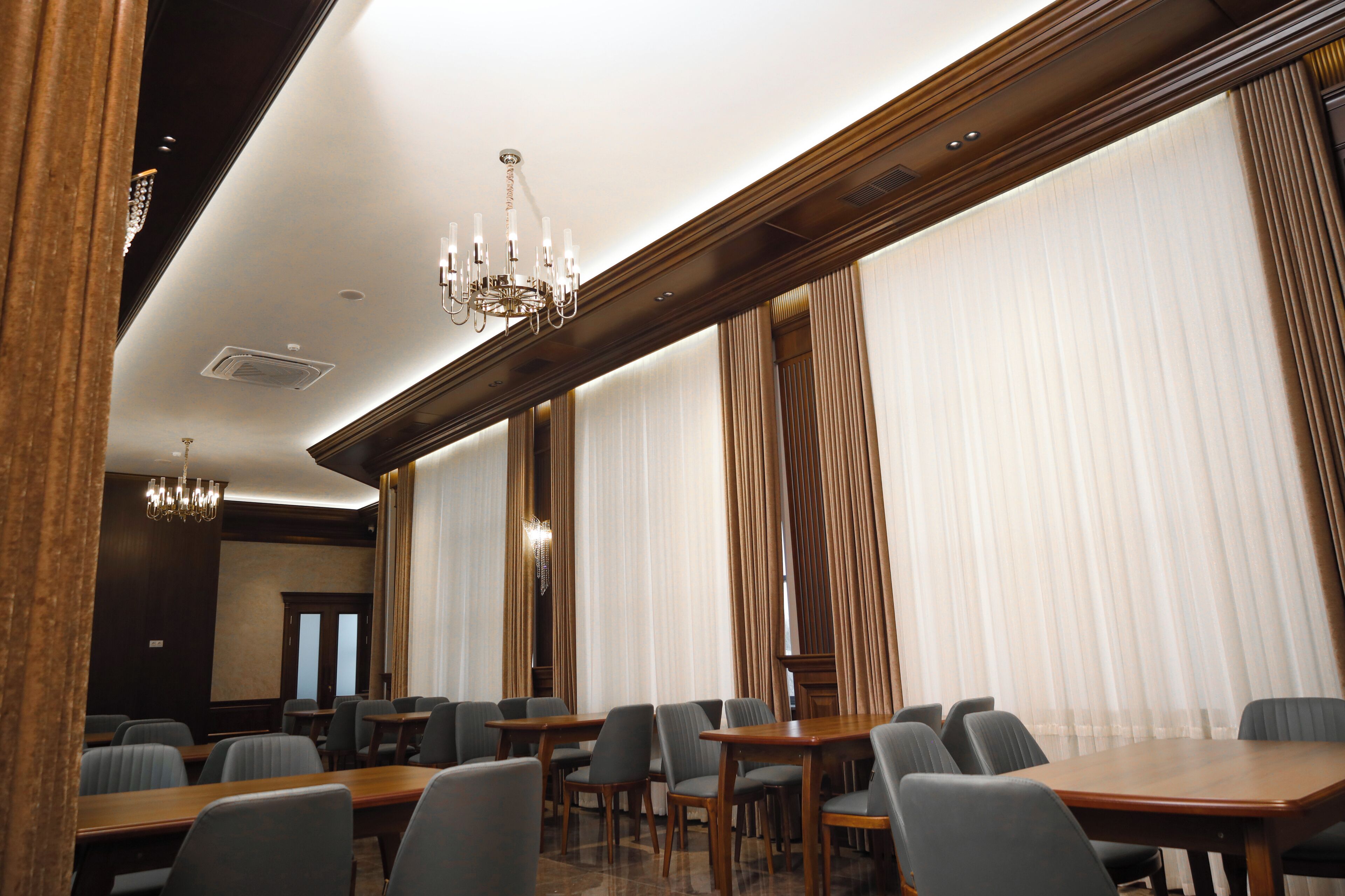 A conference room with wooden tables and chairs, large windows with curtains, and chandeliers.
