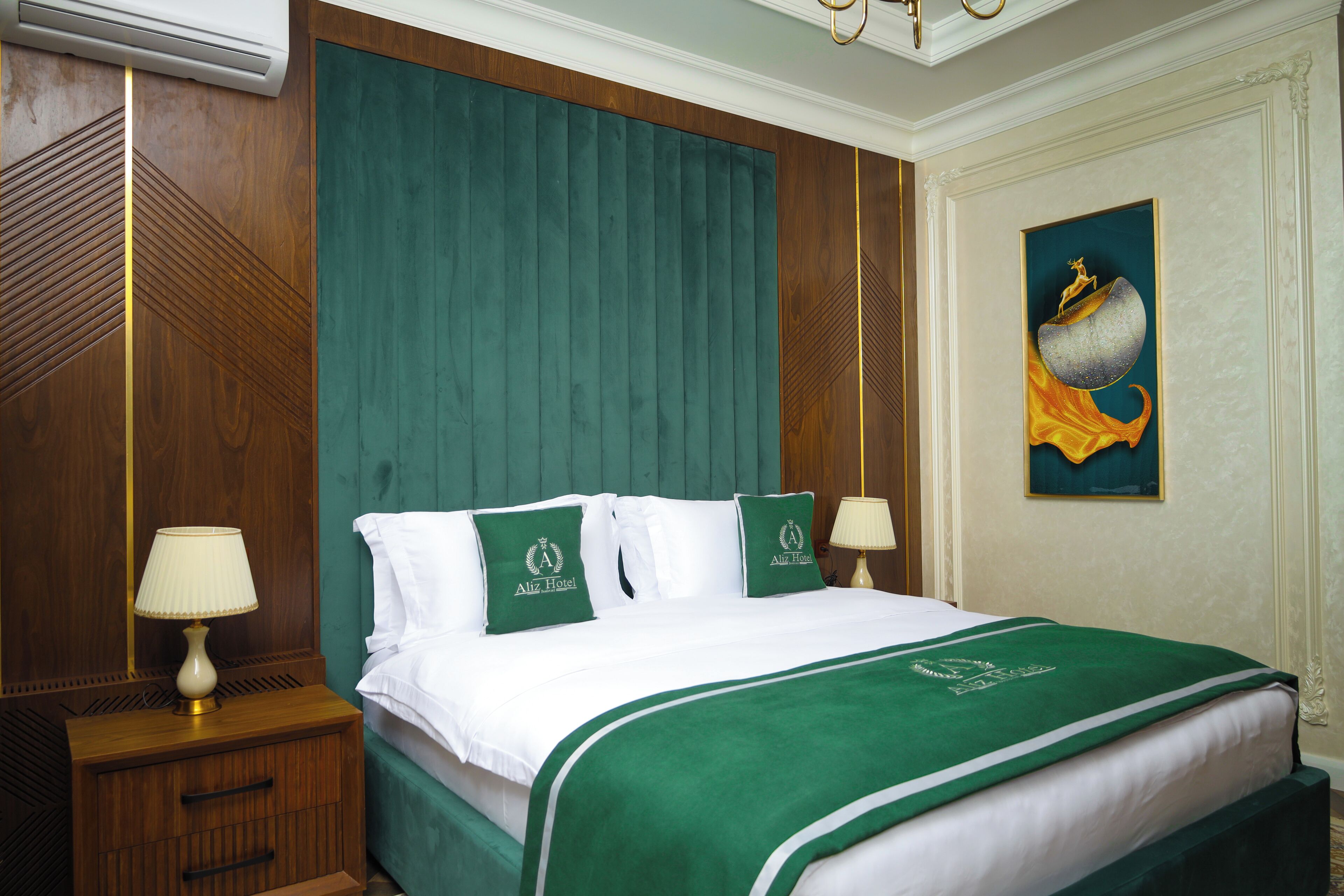 Habitación de hotel con una cama grande, ropa de cama verde, dos lámparas de noche, una mesita de noche de madera y un cuadro en la pared.