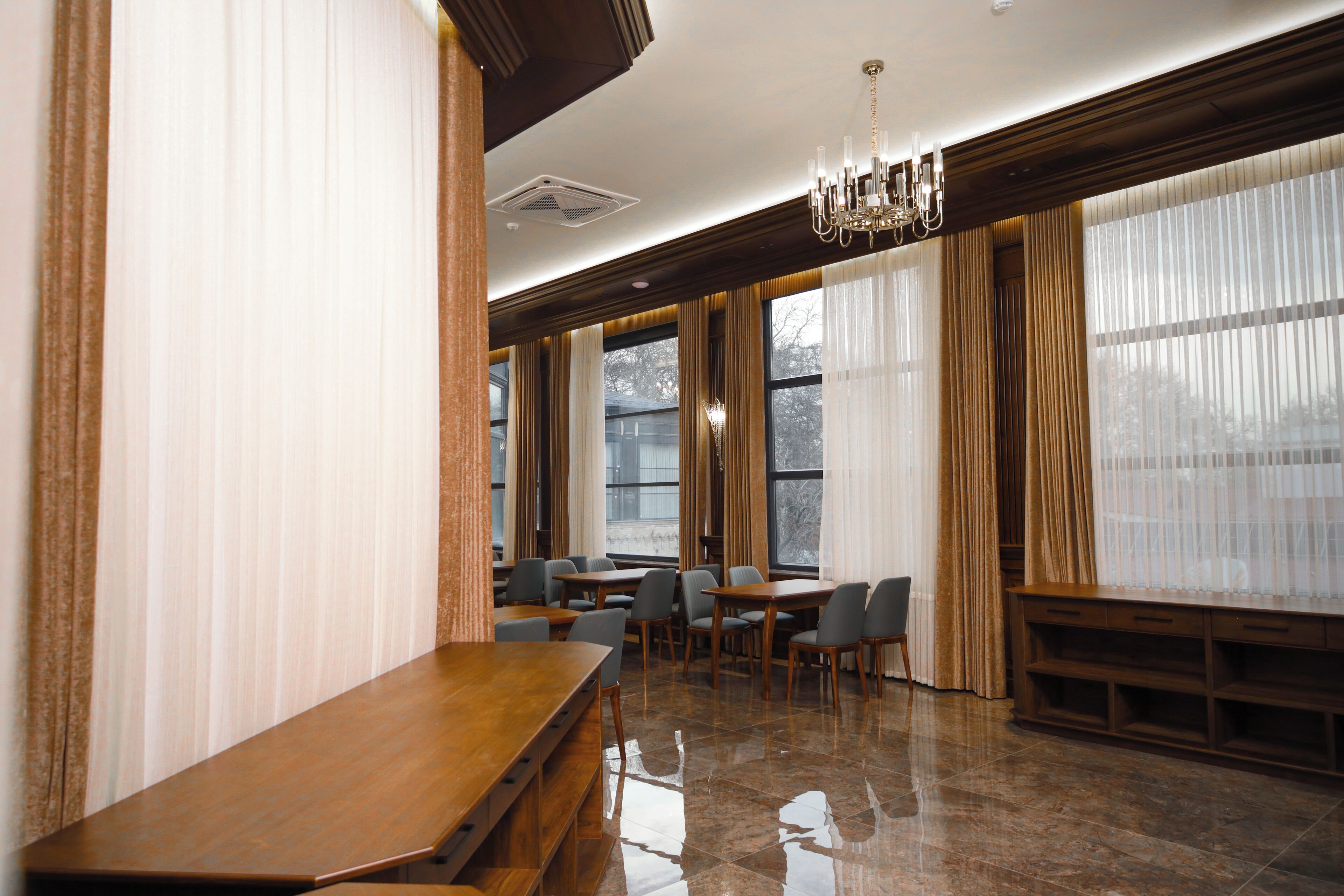 Una habitación amplia con una larga mesa de madera y numerosas sillas, ventanas grandes con cortinas y un candelabro.