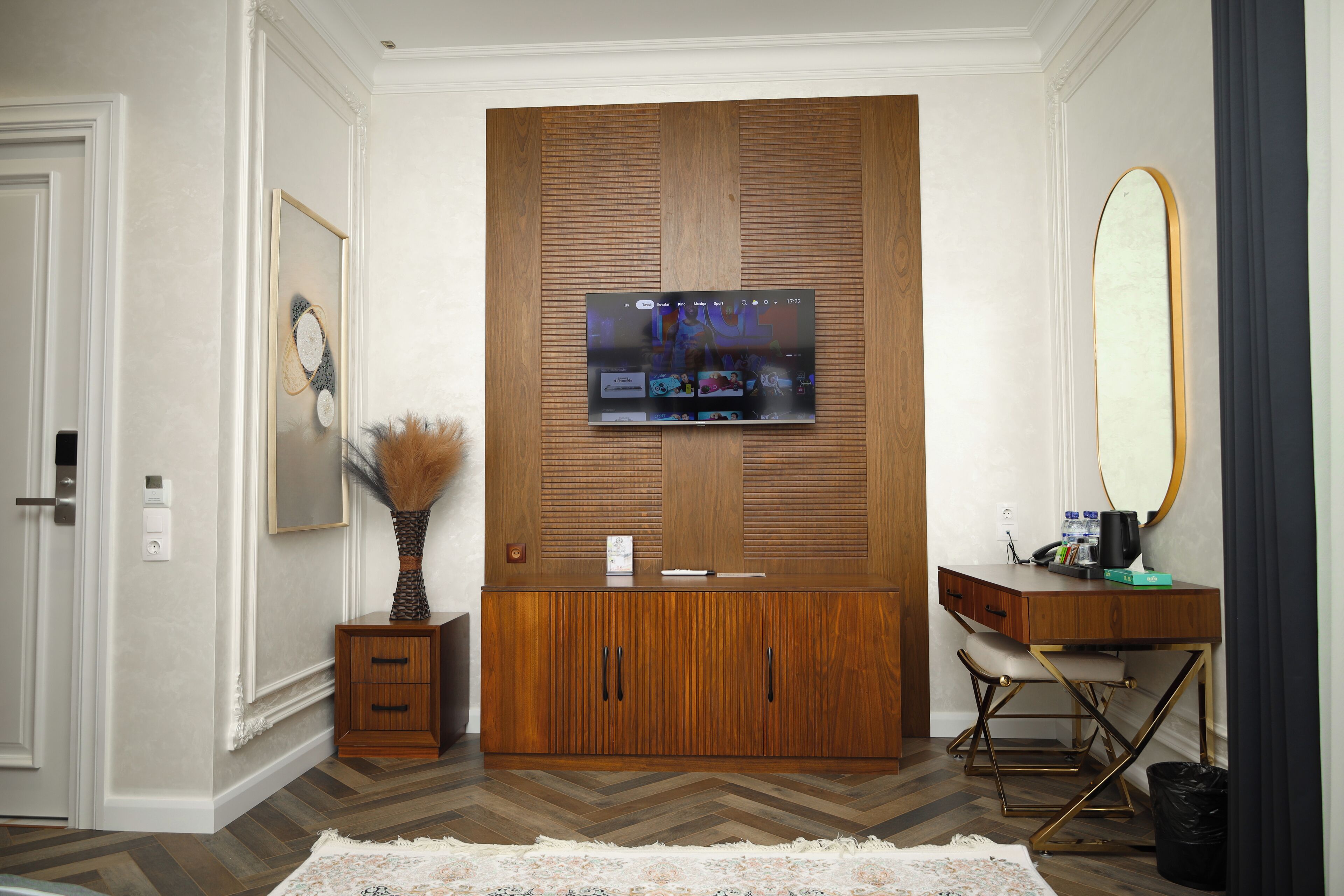 A room with a wooden wardrobe, a television mounted on the wall, a desk with a mirror, and a small side table.