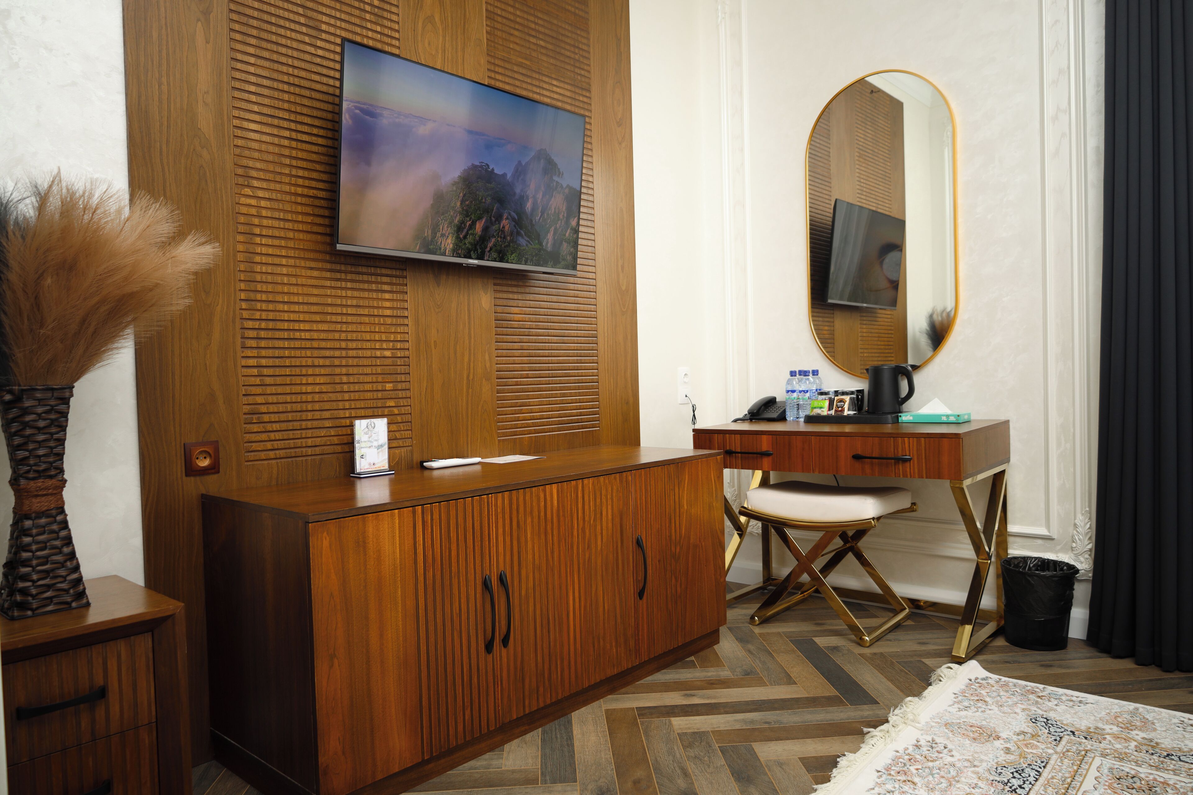 A hotel room with a wooden cabinet, a flat-screen TV, a mirror, a desk with a chair, and a patterned rug.
