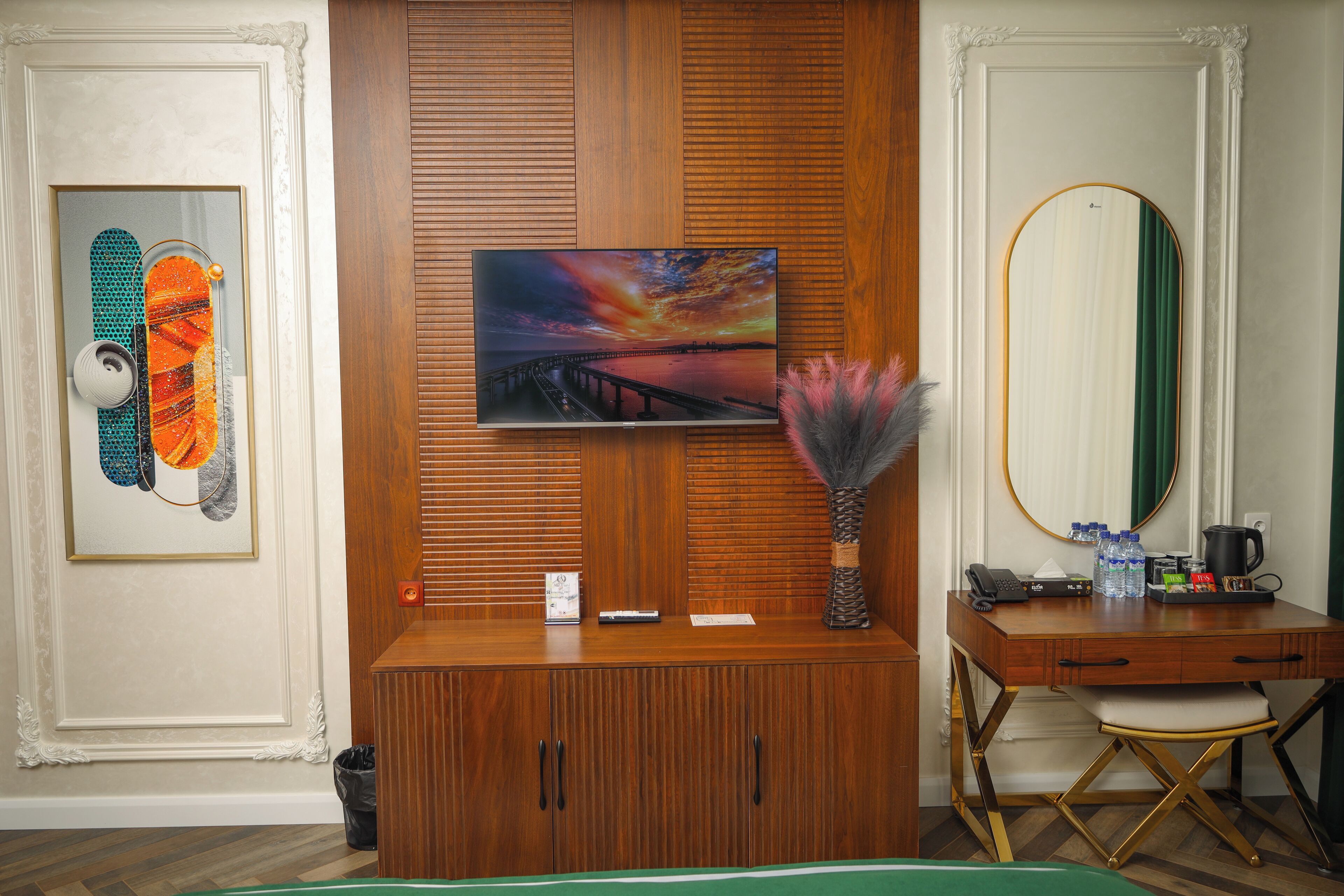 A hotel room with a wooden desk, a television mounted on a wooden panel, a framed artwork, a vase with decorative feathers, a mirror, and a green bedspread.