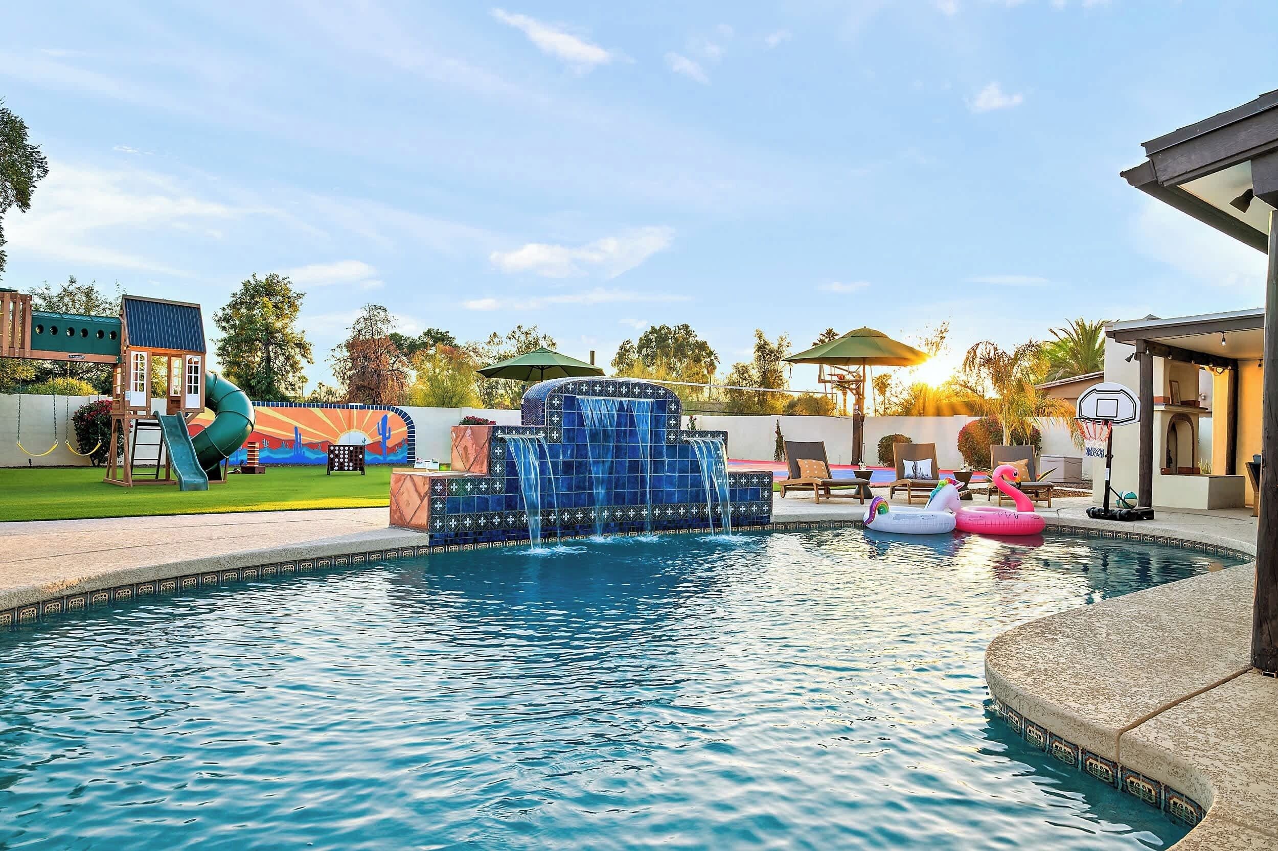 A swimming pool with a water feature, lounge chairs, and a basketball hoop.