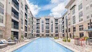 A swimming pool surrounded by multi-story buildings with balconies, lounge chairs, and outdoor seating.