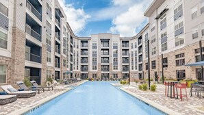 A swimming pool surrounded by multi-story buildings with balconies, lounge chairs, and outdoor seating.