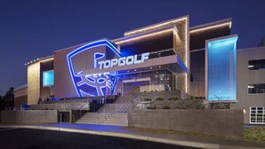 A modern building with a prominent 'TOPGOLF' sign, featuring a stylized golf club logo and a large illuminated entrance.
