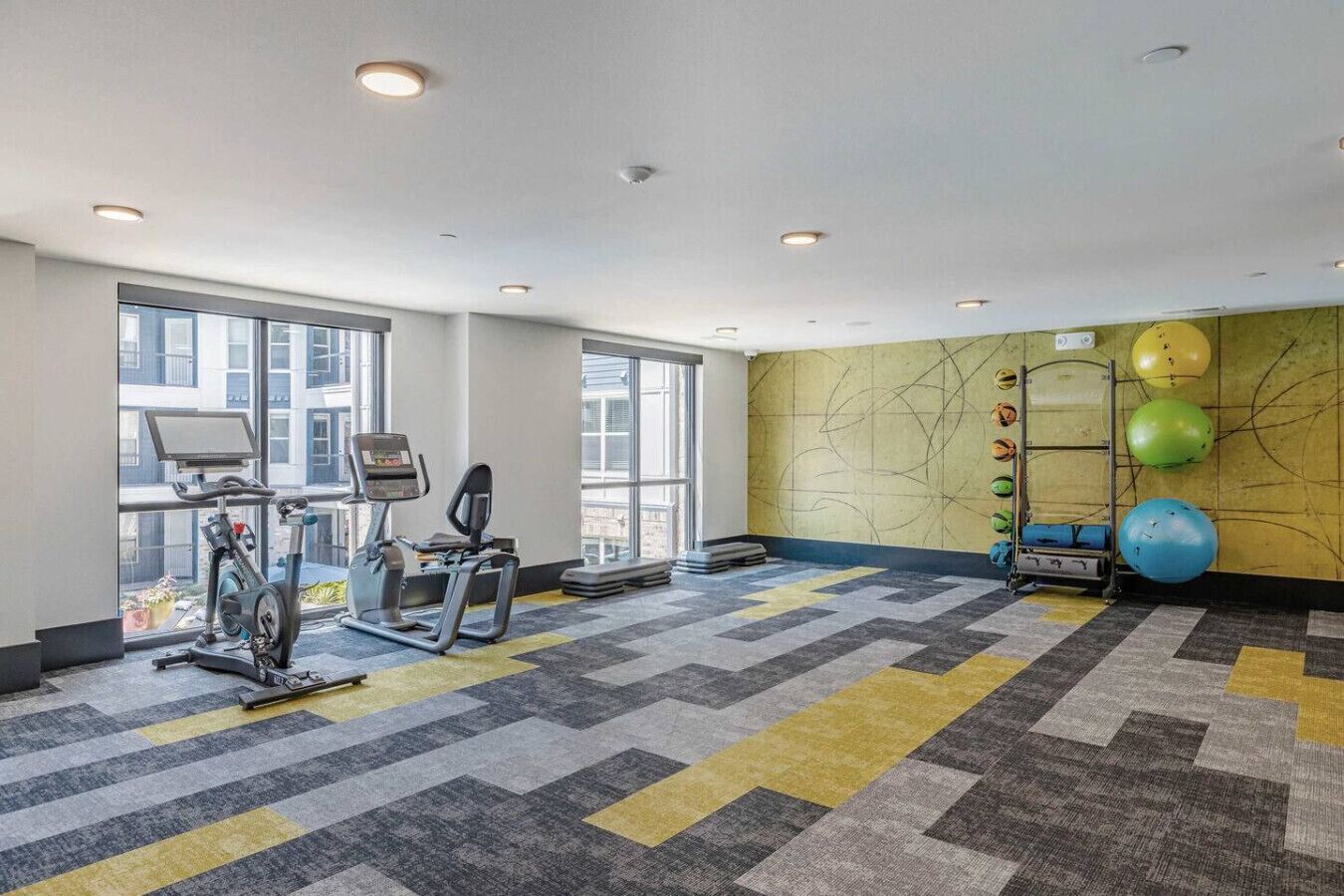 Een sportschool met trainingsapparatuur, grote ramen en een muur met fitnessballen.
