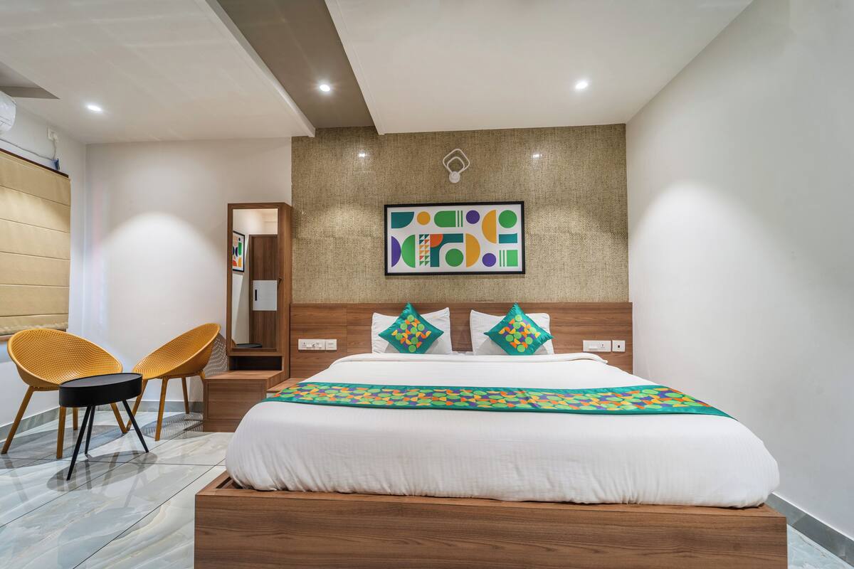 A modern hotel room with a large bed, two chairs, a wooden headboard, a wall art piece, and a small table.