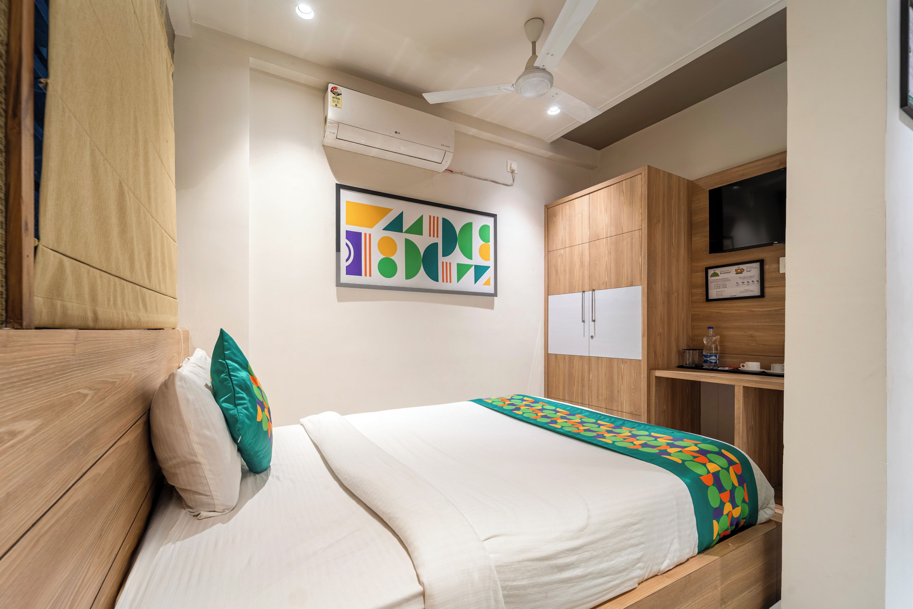 A hotel room with a bed, a ceiling fan, an air conditioning unit, a TV, and a wooden wardrobe.