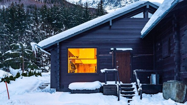 Una cabaña de madera con un ventanal grande, rodeada de árboles y montañas cubiertas de nieve.