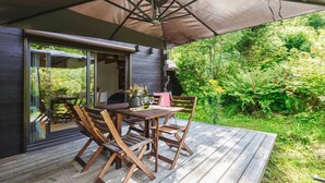 Un patio al aire libre con mesa y sillas de madera, una sombrilla grande y un ventanal de vidrio que da a un jardín.