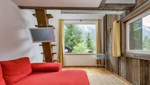 Una habitación acogedora con un sofá rojo, un televisor sobre un mueble de madera, una mesita con una computadora portátil y una ventana con vistas a árboles y montañas.