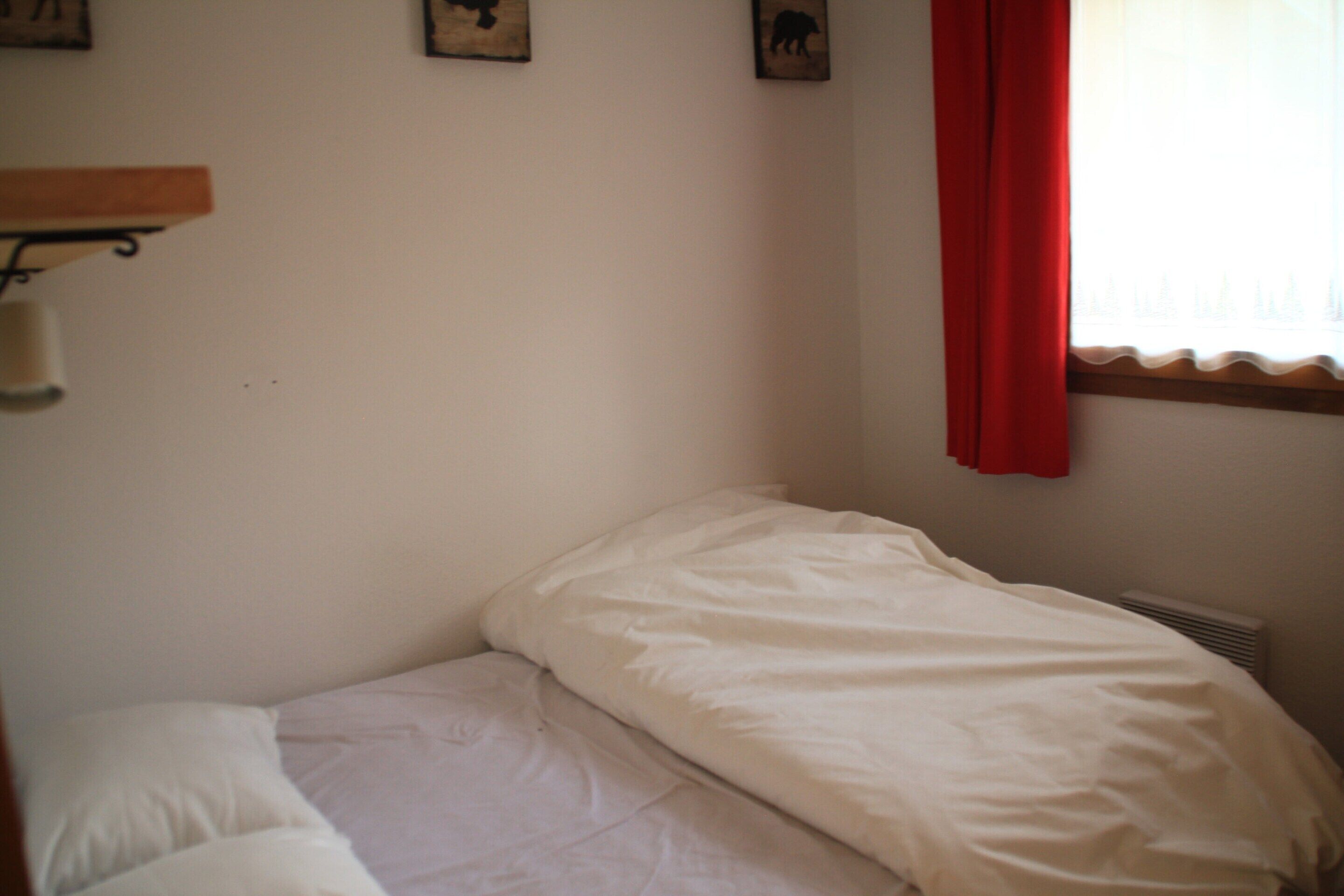 A small, single-bed room with a white bedspread, a wall-mounted shelf, and a window with a red curtain.