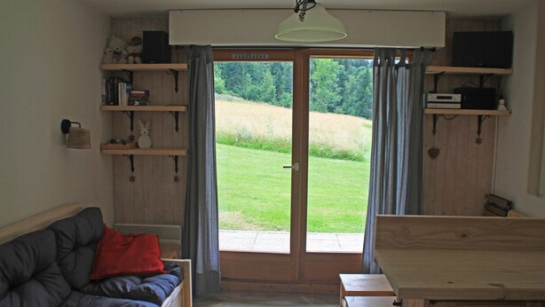 Una sala de estar acogedora con un sofá, una mesa de madera y estantes con libros y objetos decorativos. Un ventanal ofrece vistas a un campo verde.