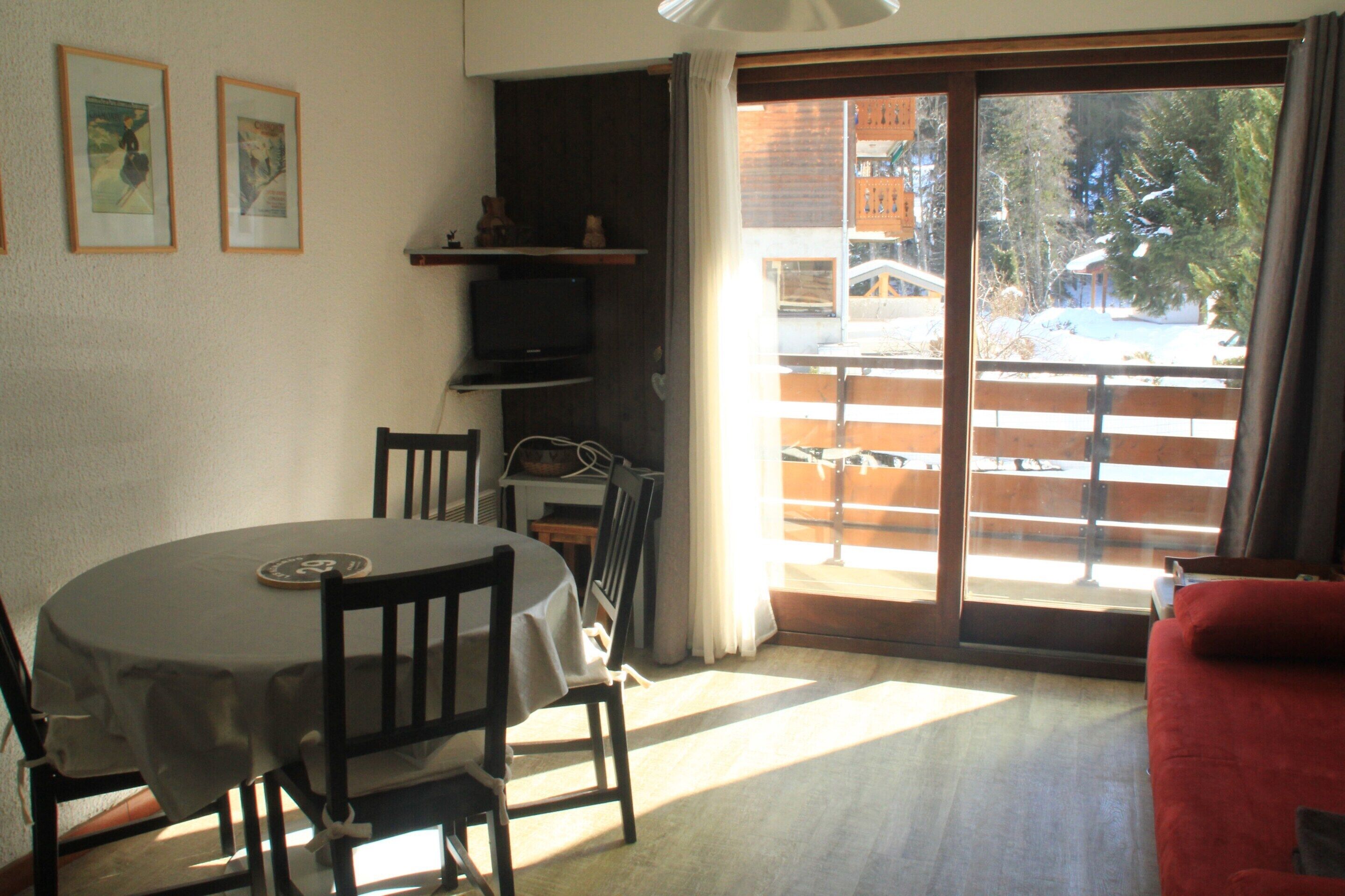 Un comedor con una mesa redonda, sillas y un televisor. Hay una puerta corrediza de vidrio que da a un balcón con vistas a un paisaje nevado.