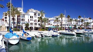 Un puerto deportivo con barcos amarrados, rodeado de edificios blancos con balcones y palmeras.