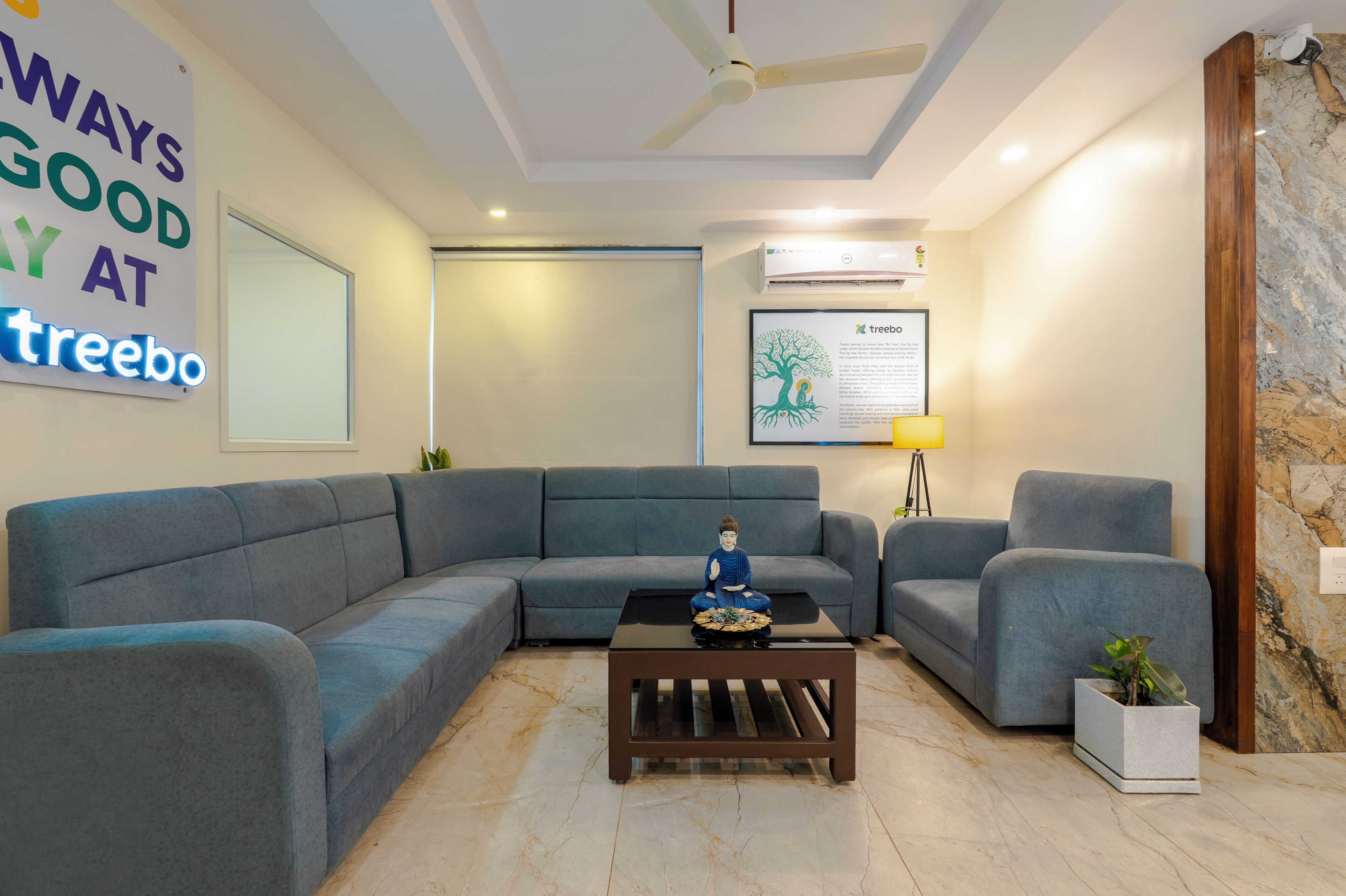 A living room with a sectional sofa, a small coffee table with a Buddha statue, a wall-mounted sign, and a framed tree illustration.