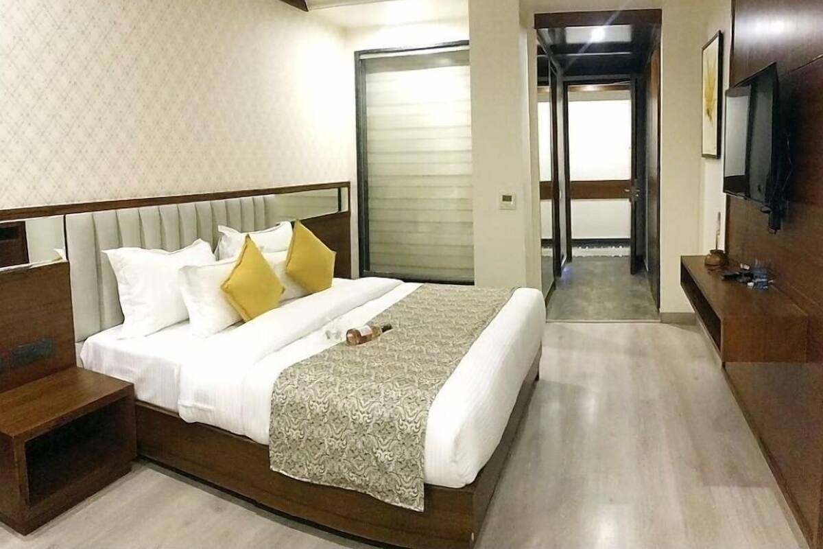 Ein Hotelzimmer mit einem großen Bett, einem Holfnachtkästchen, einem an der Wand montierten Fernseher und einer Tür, die in einen weiteren Raum führt.