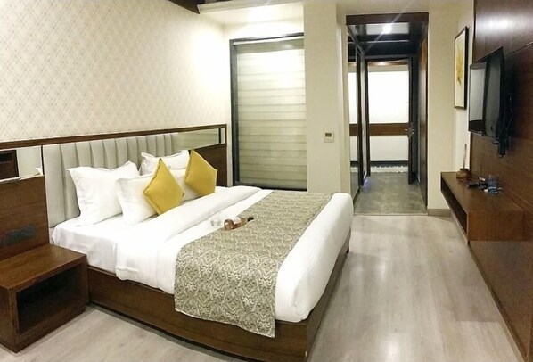Habitación de hotel con una cama grande, una mesita de noche de madera, un televisor montado en la pared y una puerta que da a otra habitación.