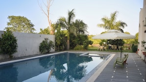 Una piscina rectangular con borde de azulejos, rodeada por un patio con una sombrilla blanca y sillas verdes.