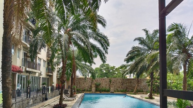 A swimming pool surrounded by palm trees and a stone wall, with a building in the background. - Amerta Room Apartemen Mutiara by Cocotel (Bekasi)