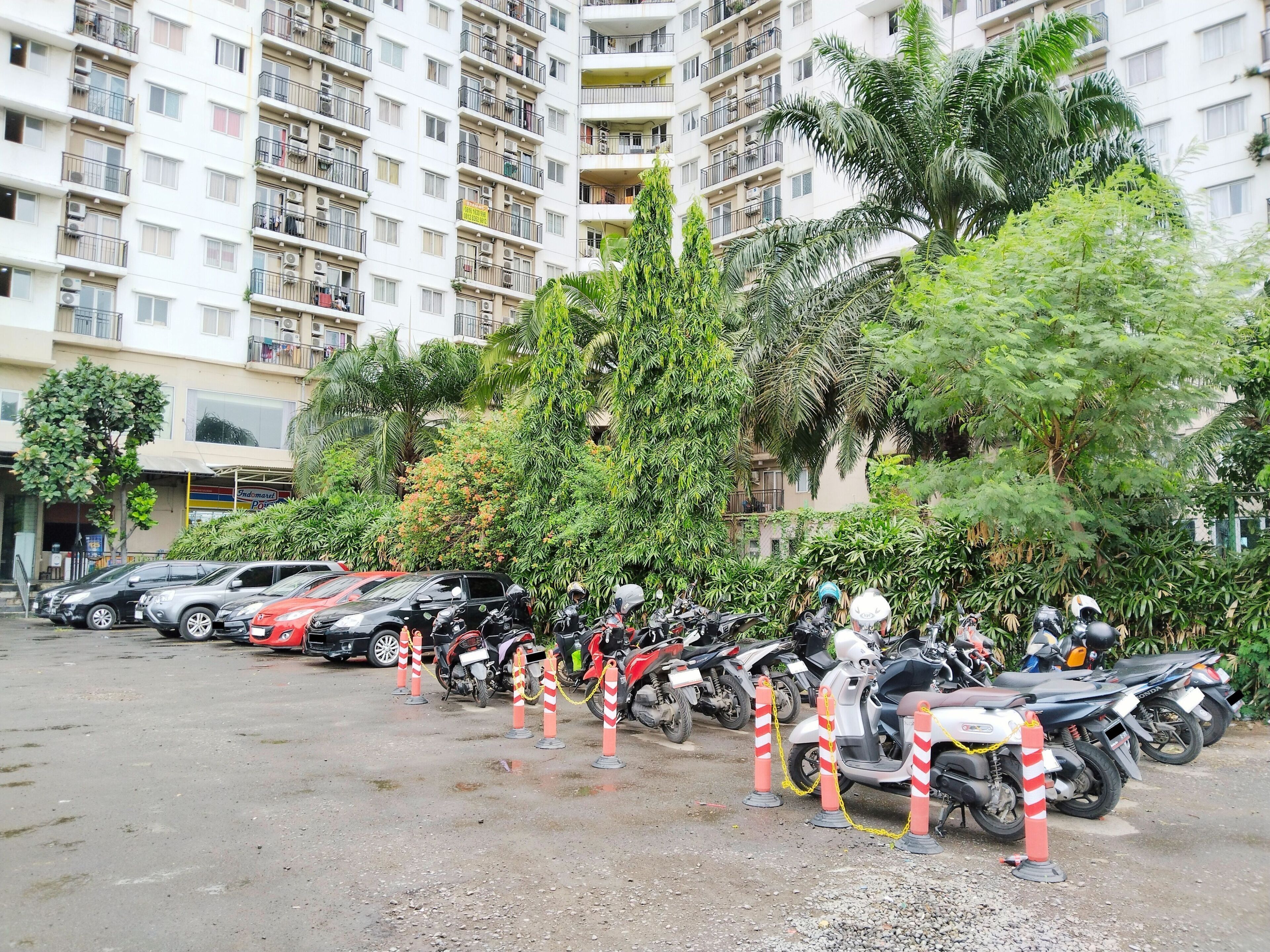 A parking area with multiple motorcycles and cars, surrounded by greenery and residential buildings.
