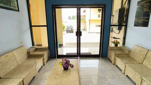 Uma área de recepção com balcão, dois sofás bege, uma mesinha com um vaso de flores e uma porta de vidro que dá para o exterior.