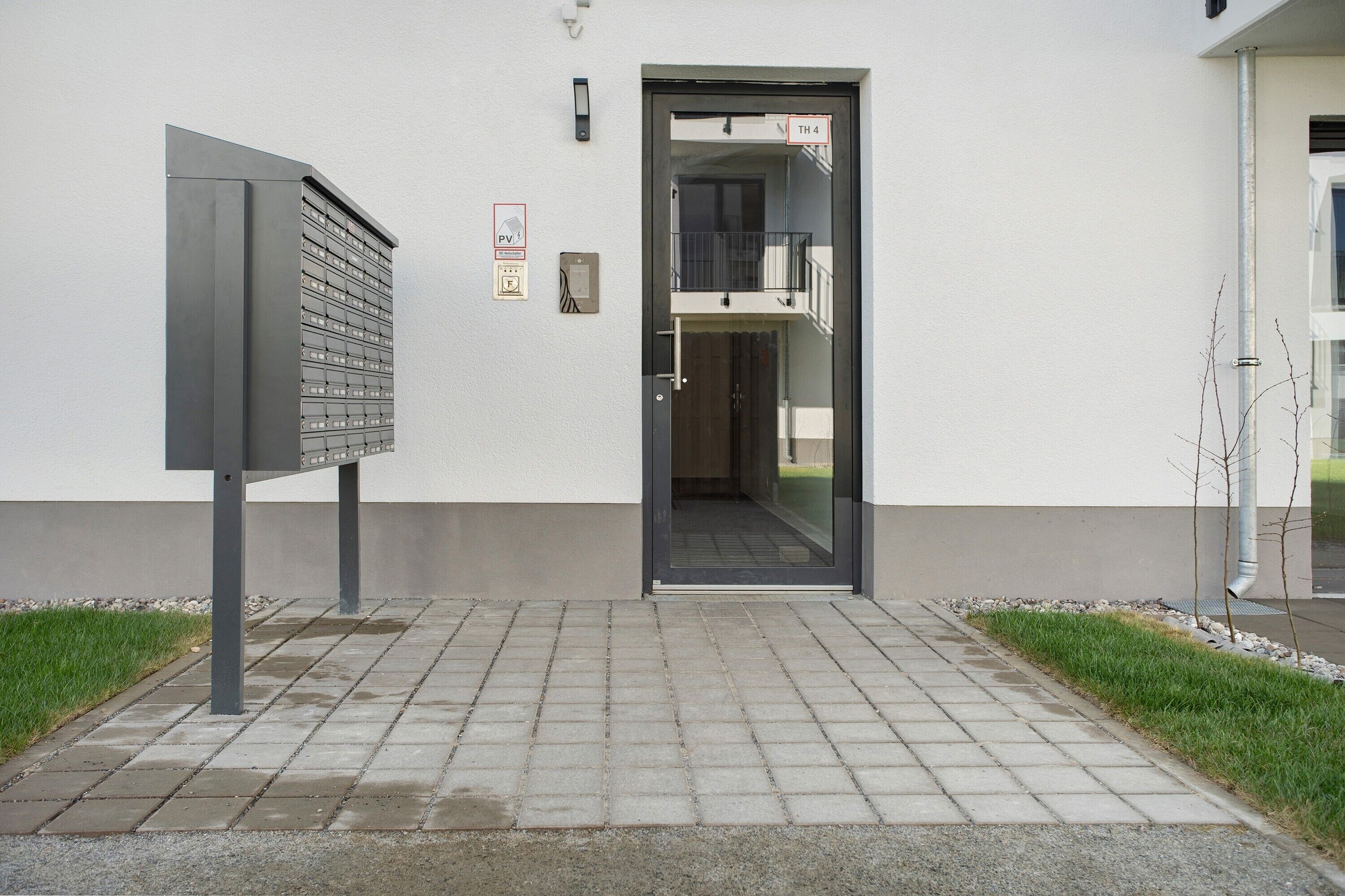 Entrada de un edificio con un buzón, una puerta con la etiqueta 'TH 4' y un pequeño espacio exterior con césped y un sendero.