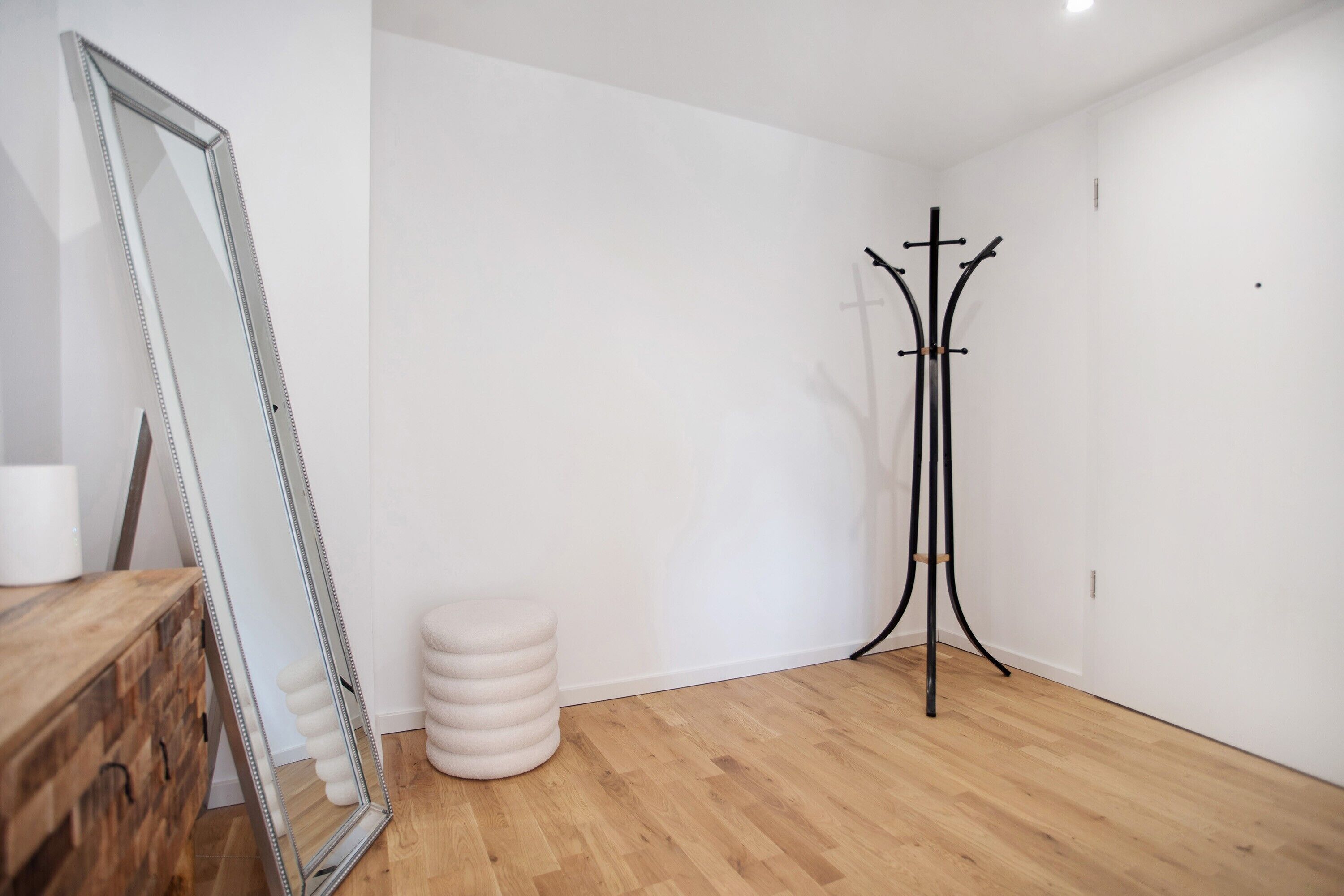 Una habitación con suelo de madera, un espejo, un taburete redondo, un perchero y un aparador de madera.