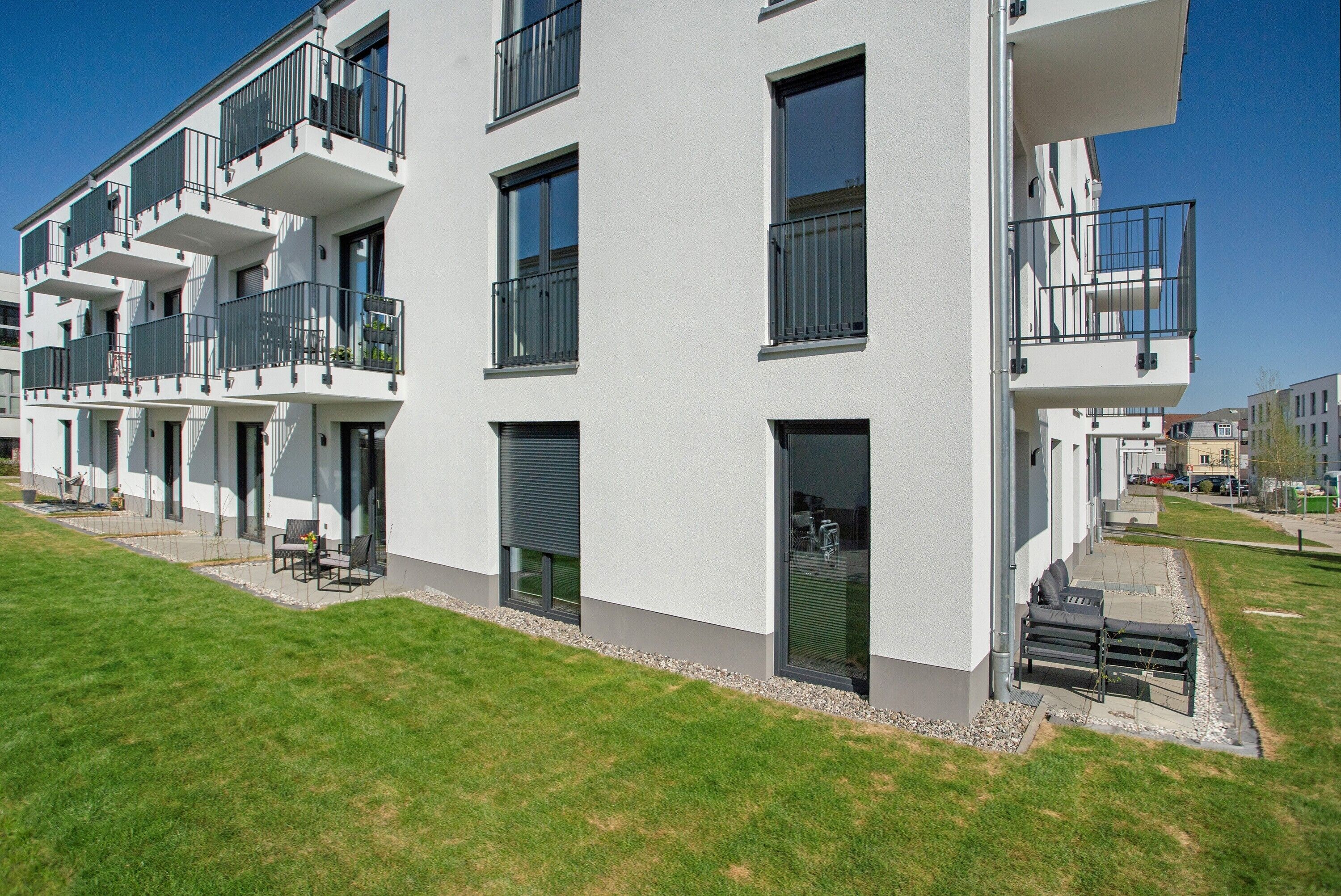 A multi-story residential building with balconies, a grassy lawn, and a clear sky.