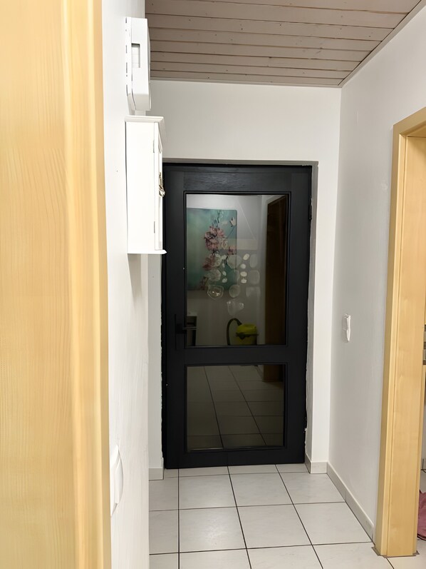 A hallway with a dark door, a white wall, a light switch, and a tiled floor.
