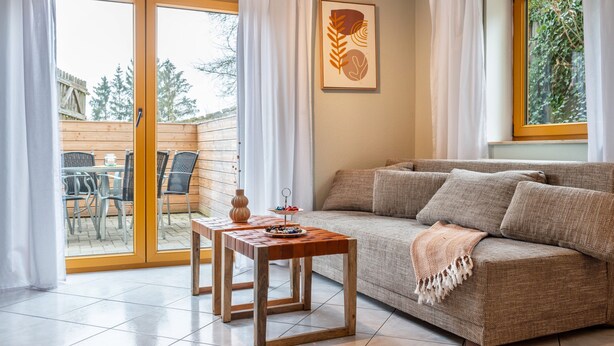 A living room with a sofa, a wooden table, and a glass door leading to an outdoor area with chairs and a wooden fence. - Apartment 'Donauliebe' with Private Terrace, Shared Garden and Wi-Fi (Sigmaringen)