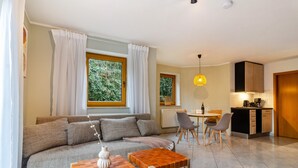 A modern living room with a sofa, two wooden tables, a dining area with chairs, a kitchen with cabinets, and large windows with curtains. - Apartment 'Donauliebe' with Private Terrace, Shared Garden and Wi-Fi (Sigmaringen)