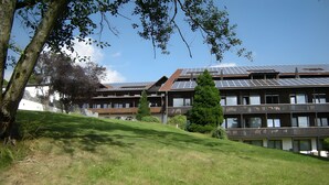 Een meerhoog gebouw met zonnepanelen op het dak, een grashelling en een boom in de voorgrond.