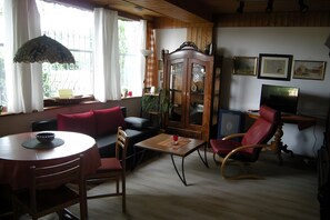 Uma sala de estar com uma mesa de jantar de madeira, um sofá, uma poltrona vermelha, um armário e uma televisão.