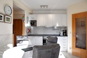 A compact kitchen with white cabinets, a microwave, and a stove. There is a dining table with a chair in the foreground. - Apartment 'Tannenblick 8-1' with Private Terrace, Shared Garden and Wi-Fi (Oberhof)