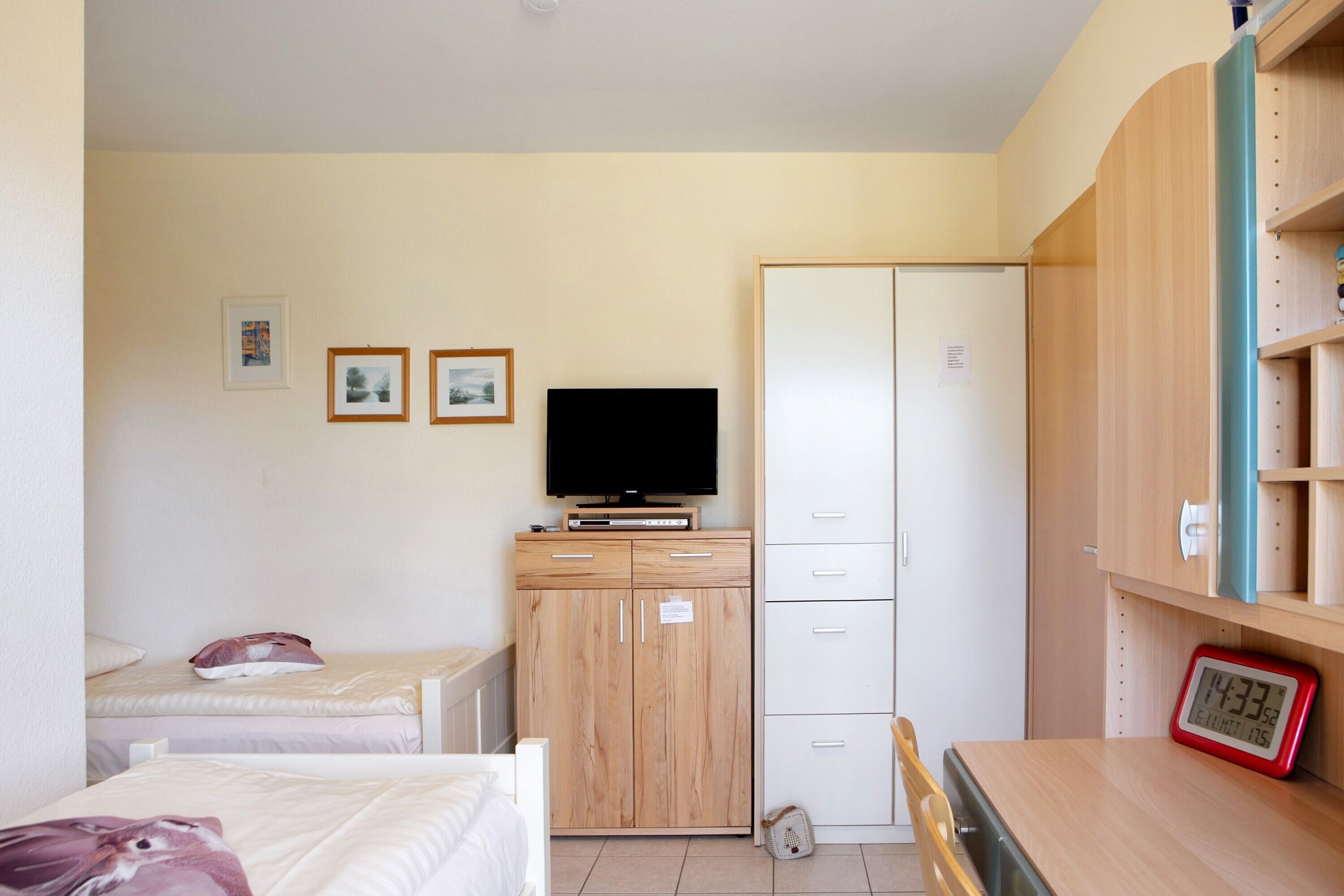 Une chambre à coucher comprenant un lit, une télévision, une armoire, un bureau avec une horloge et une petite étagère.