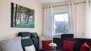 Un saló amb un sofà fosc, coixinets vermells i blancs, un quadre d'arbres i una finestra amb cortines.