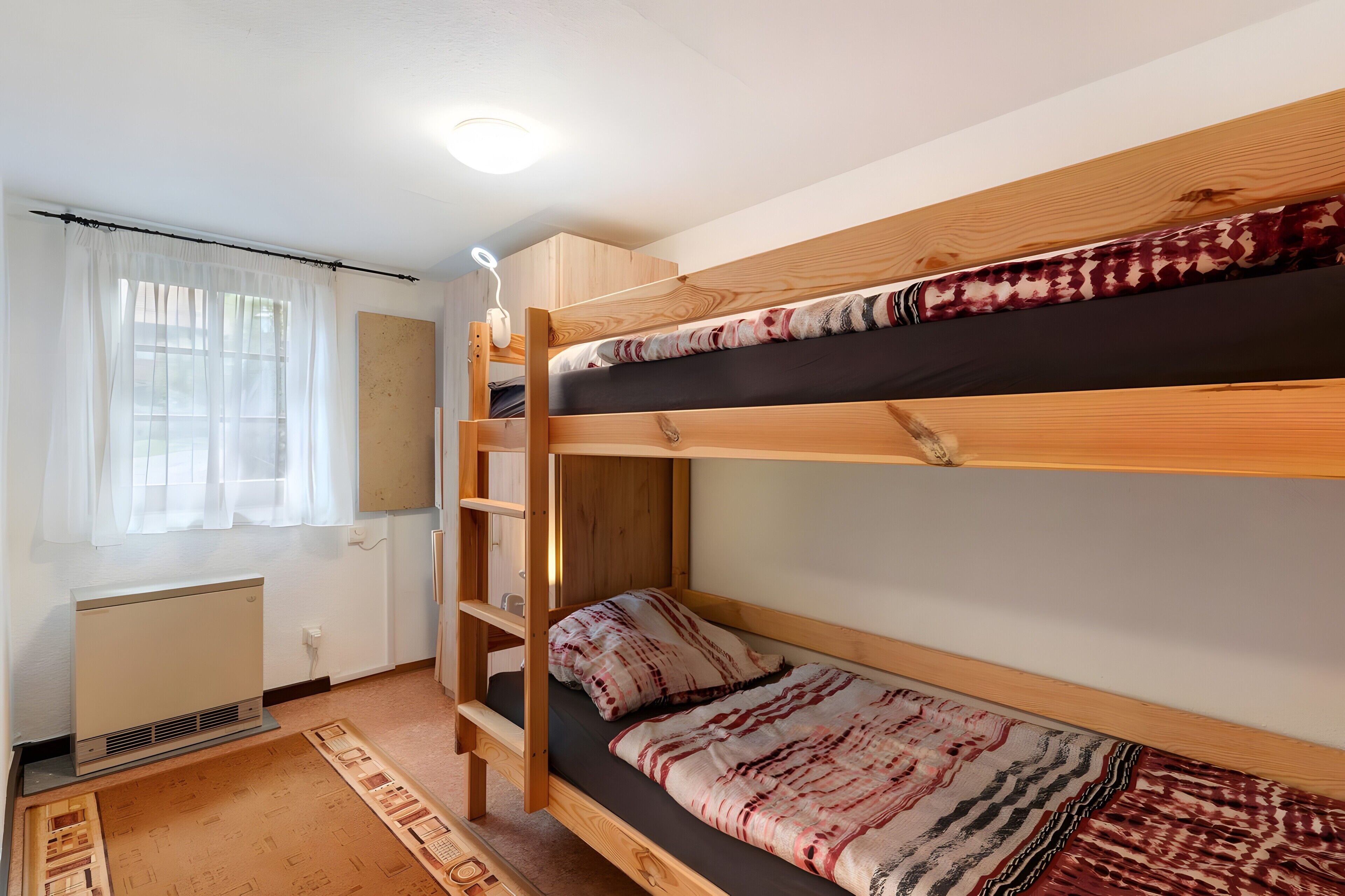 Une chambre avec un lit superposé doté d’une tête de lit en bois, un petit bureau avec une lampe, un radiateur et un tapis à motifs.