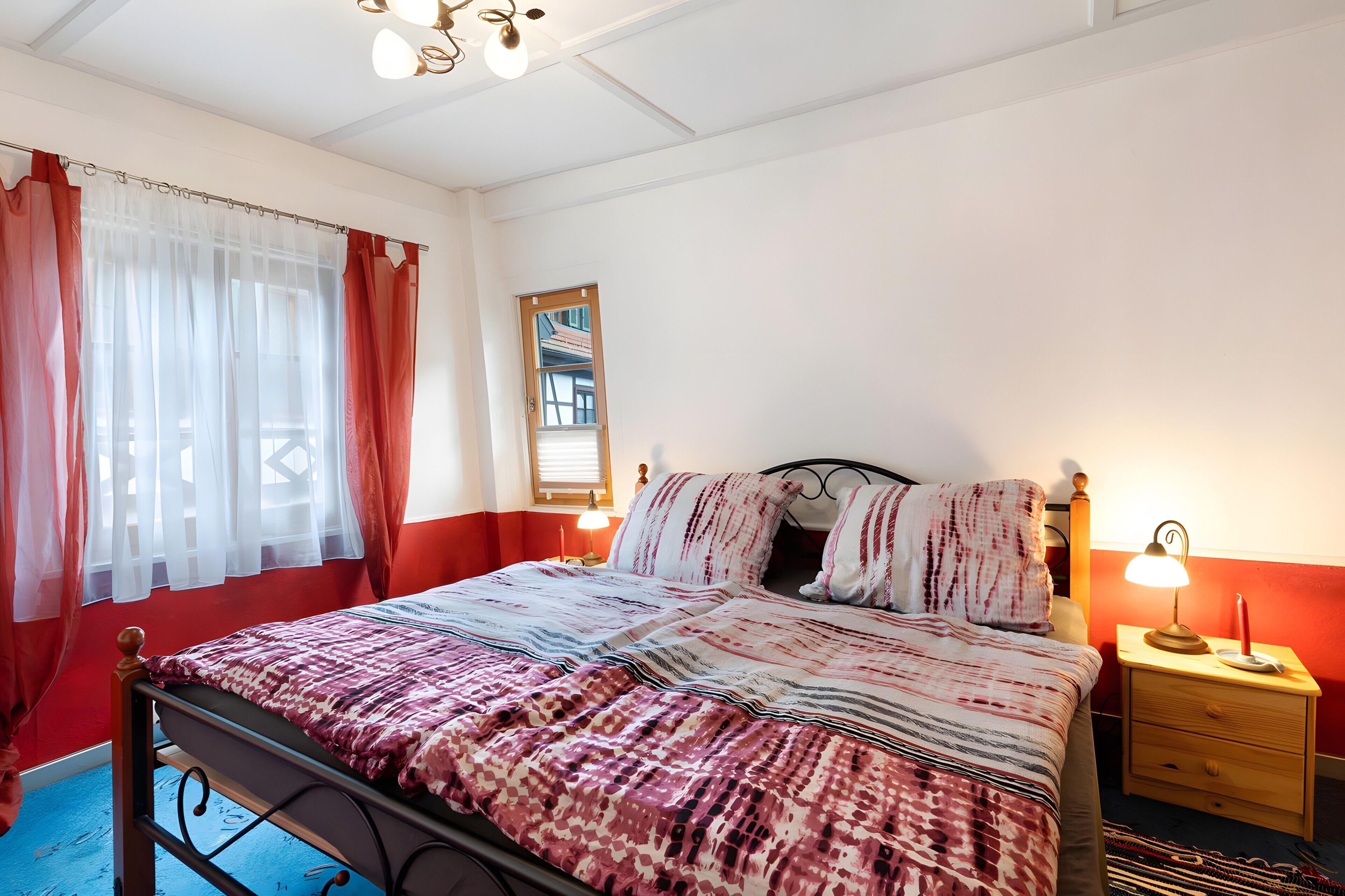 Ein Schlafzimmer mit Bett, Nachttisch, Lampe, einem Fenster mit Vorhängen und einem Spiegel an der Wand.