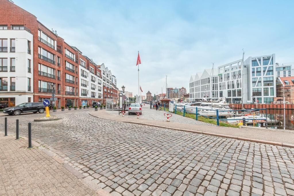 A cobblestone street with modern buildings, a flag, and boats docked by the water.
