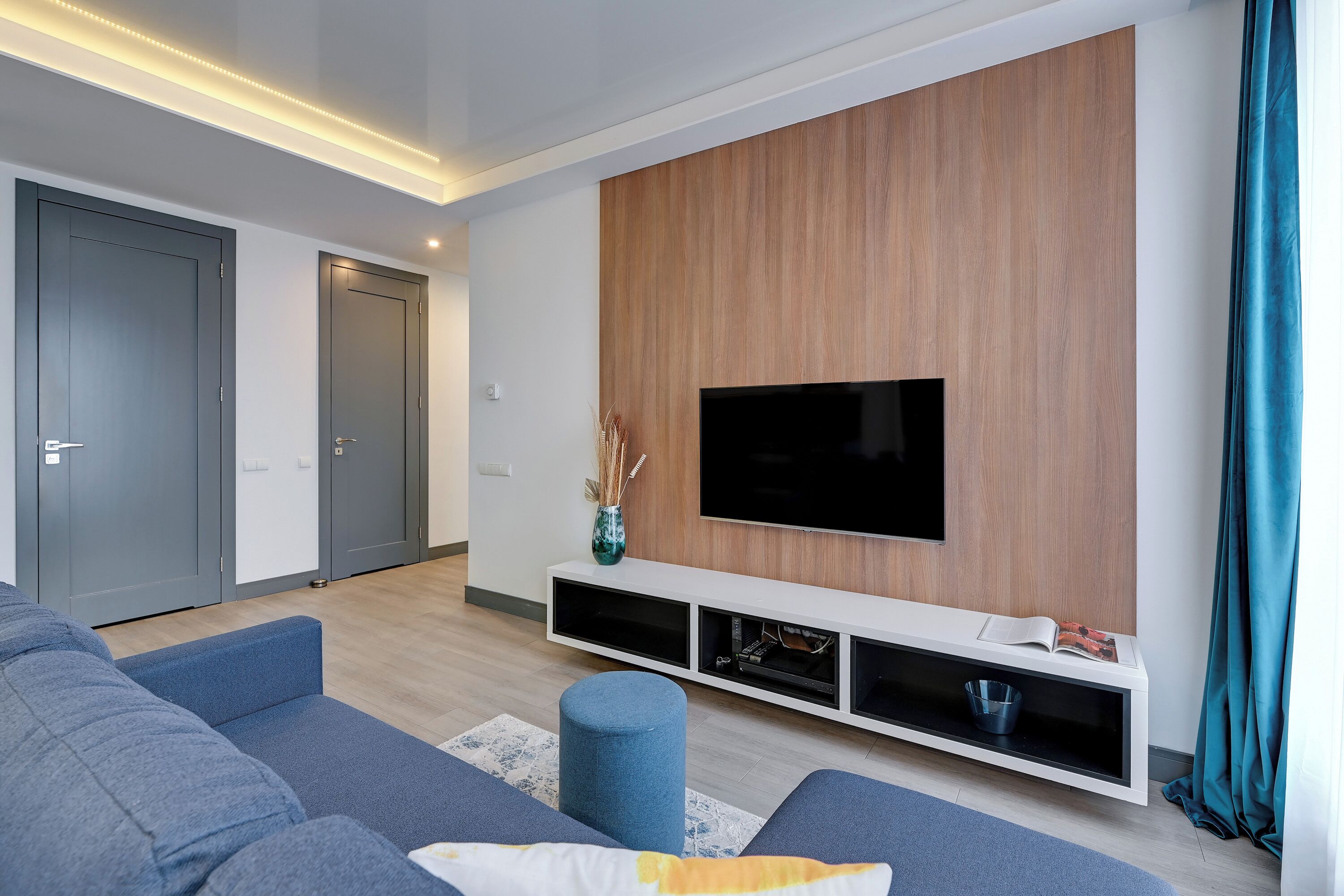 A modern living room with a flat-screen TV mounted on a wooden wall, grey sofas, a blue ottoman, and a white entertainment unit.