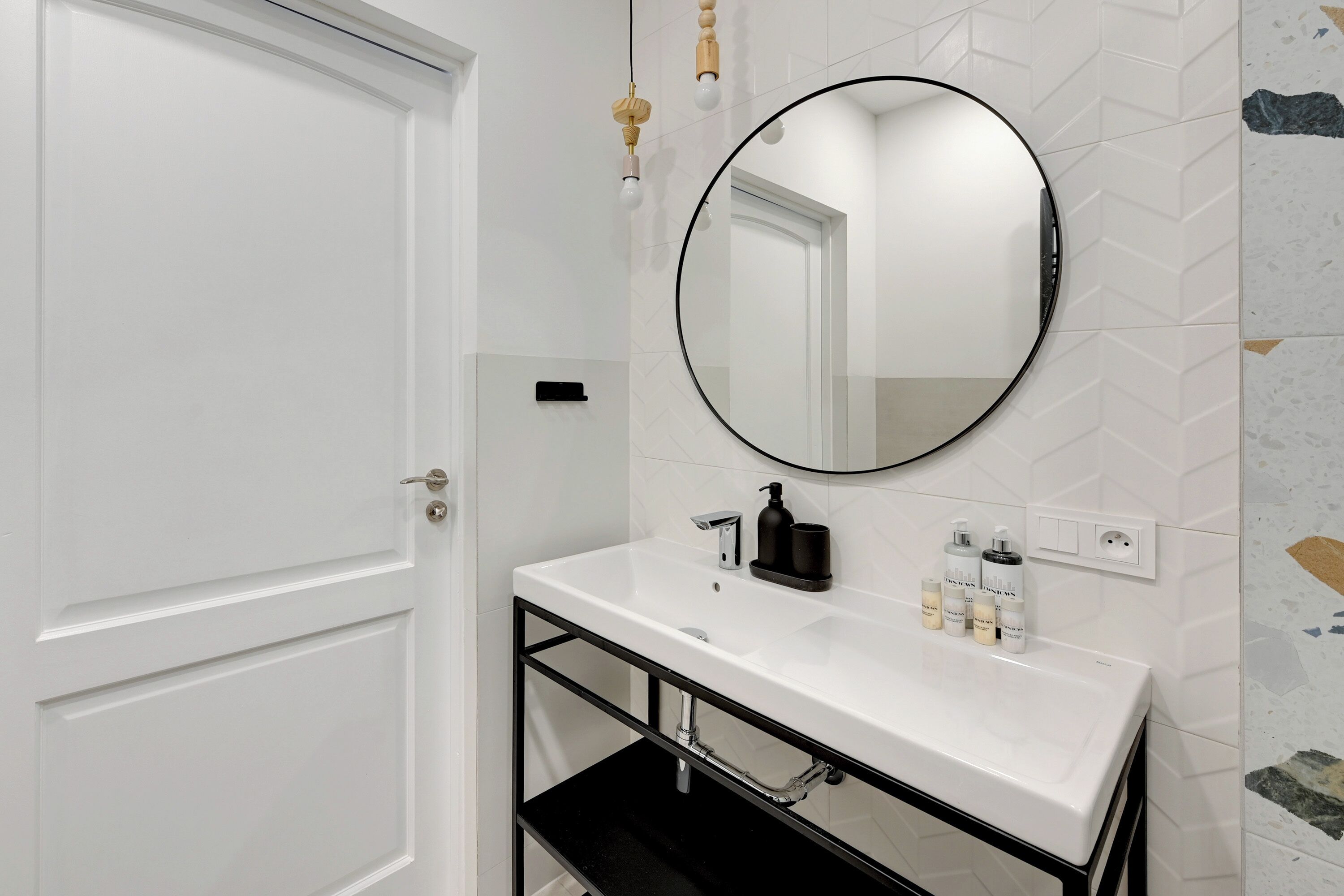 A bathroom with a white sink, black faucet, and a round mirror. There are toiletries on the sink and a towel rack above it.