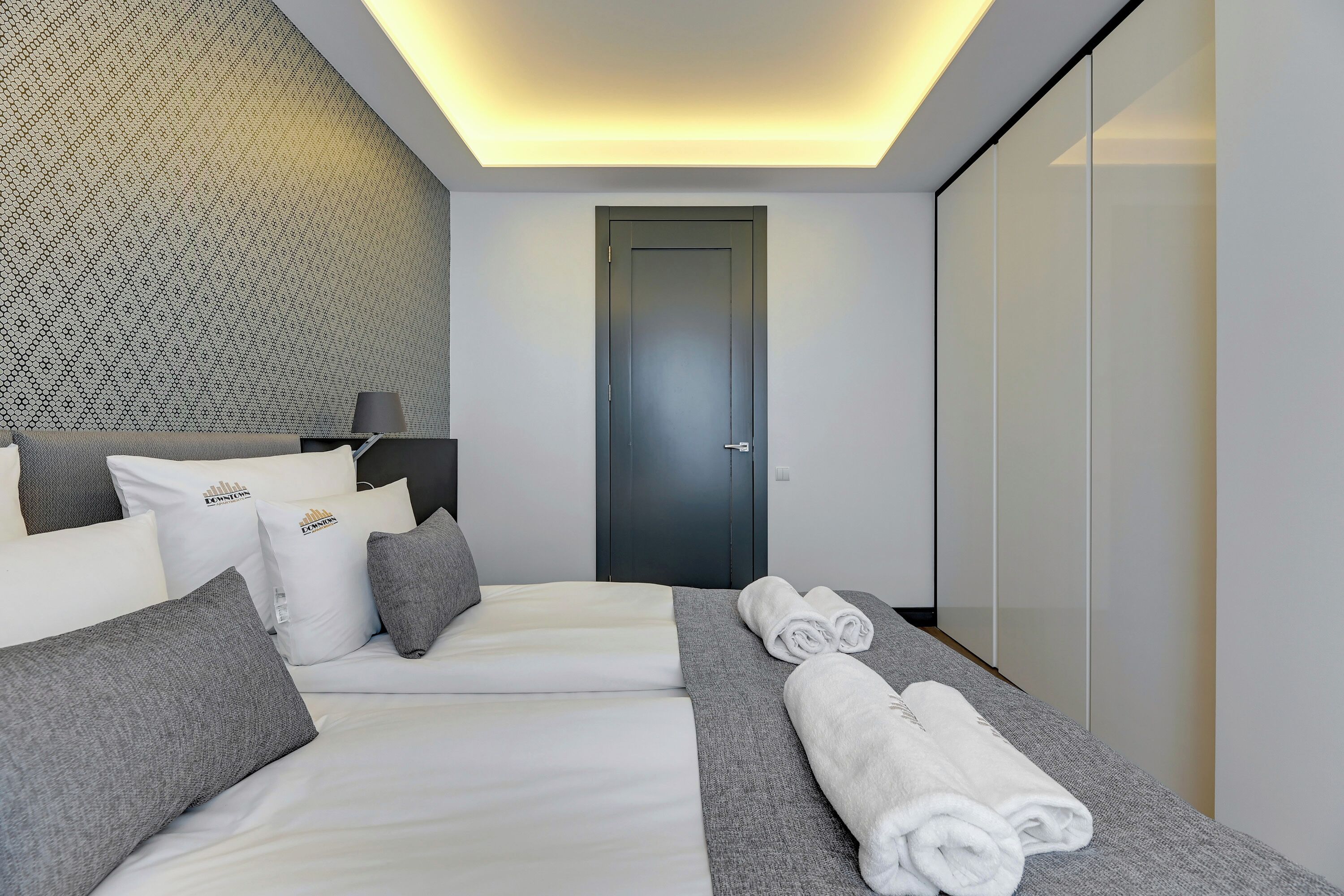Moderni hotellihuone, jossa on siististi pedattu sänky, harmaan ja valkoisen värimaailma sekä kuvioitu seinä.