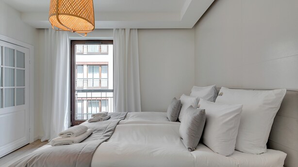 A bedroom with a large bed, white bedding, a hanging light fixture, and a view of a building through a window. - Riverside Szafarnia Street by Downtown Apartments (Gdansk)